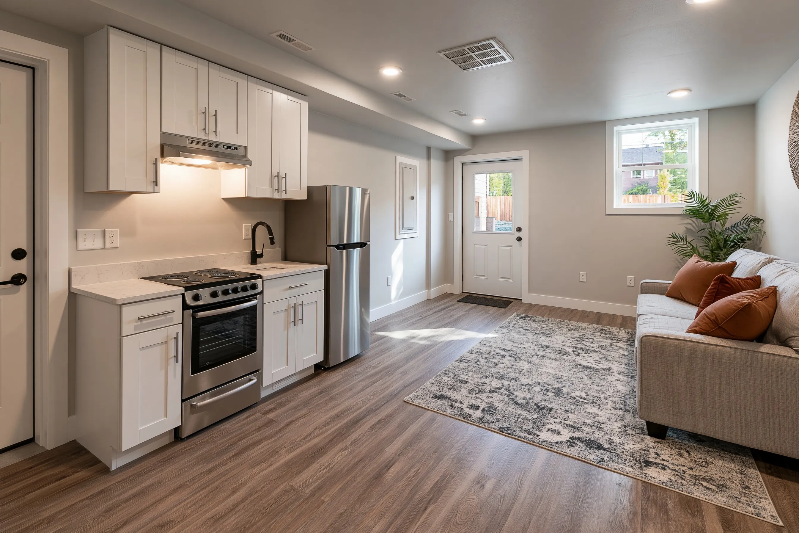 Basement apartment main room with a kitchenette along one wall, shaker cabinets, a small range and refrigerator, a loveseat and a separate exterior door with a glass panel