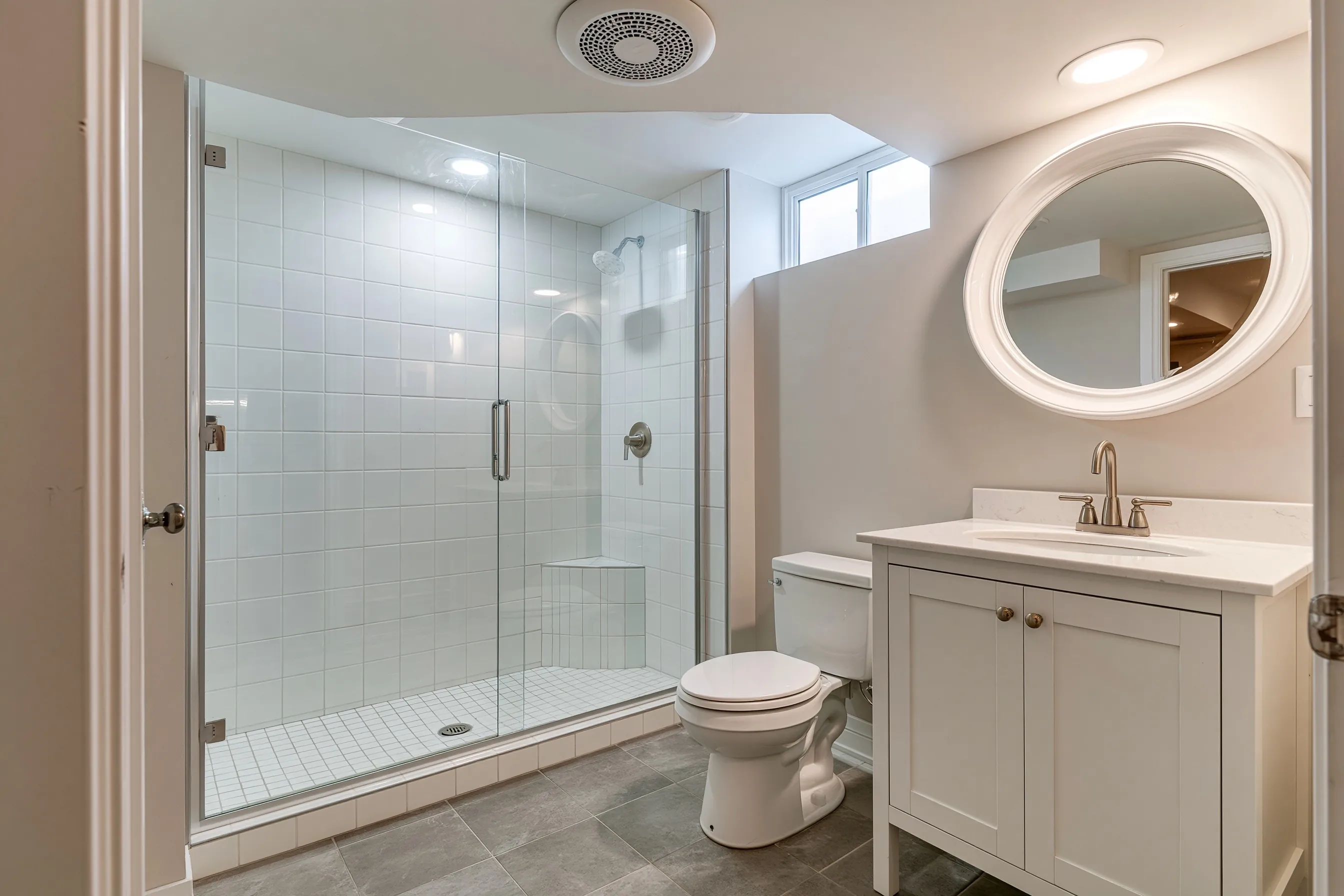 Finished basement bathroom with a tiled walk-in shower and glass panel, a white vanity, grey porcelain tile floor, an exhaust fan grille and one small window